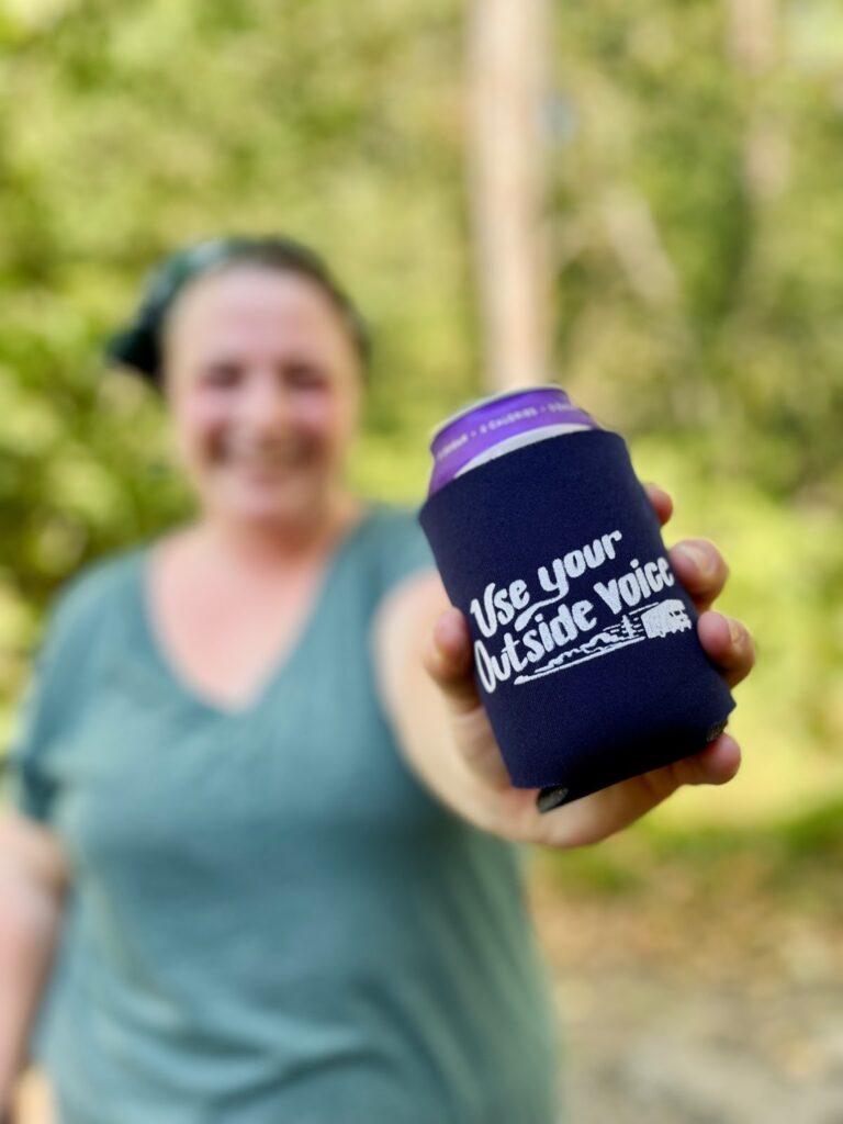 Woman holds out a can in a coozy that reads 'use your outside voice'